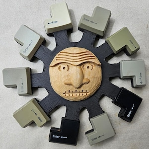 May include: A unique wall art piece featuring a carved wooden face at the center, surrounded by repurposed computer "Enter" keys. The keys are in various shades of gray and black, arranged in a circular pattern around the face.