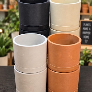 May include: A collection of six cylindrical plant pots arranged in two stacks of three. The pots feature a smooth, matte finish and come in four distinct colors: black, off-white, light gray, and terracotta orange, each with a matching base.