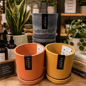 May include: Three cylindrical plant pots with matching saucers. One pot is a dark gray with a textured, cloudy pattern, while the others are solid orange and yellow. Each pot features a vertical black label with the white text KANSO DESIGNS.
