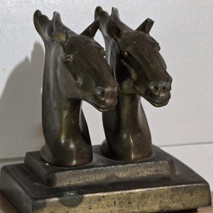 Peut inclure: Sculpture en bronze représentant deux têtes de chevaux côte à côte. La sculpture présente une patine sombre et vieillie, posée sur un socle rectangulaire. Les têtes de chevaux sont détaillées, avec des yeux, des oreilles et des traits du visage, représentant des formes équines.