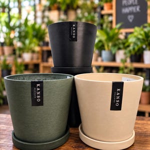 May include: Three tapered plant pots with matching saucers in black, olive green, and cream. Each pot features a vertical black label with the white text KANSO DESIGNS. The pots have a textured finish and are arranged in a tiered formation.