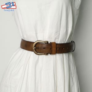 May include: A brown leather belt with a brass buckle. The belt has a distressed look and is worn on a white dress.