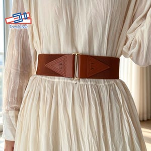 Peut inclure: Une large ceinture élastique marron dotée de deux empiècements triangulaires en similicuir au centre. Les empiècements sont gaufrés avec les lettres GK et reliés par un fermoir à crochet en métal doré. La ceinture est portée sur une robe crème.