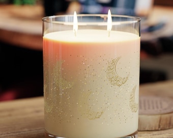 May include: A clear glass candle jar with a white and gold candle inside. The candle has a pink and white ombre effect and a design of gold moons and stars. The candle is lit with two wicks.