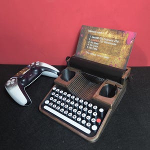 Puede incluir: Un soporte para mando de videojuegos de estilo máquina de escribir marrón y negro con un mando de videojuegos blanco y negro y una tarjeta con texto. El soporte tiene un teclado con teclas blancas y negras y una ranura para la tarjeta. La tarjeta tiene el texto "Memory card".