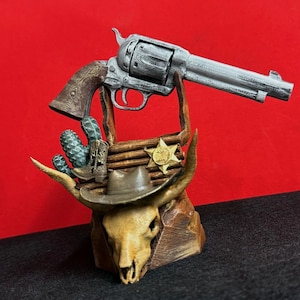 May include: A decorative Western-themed display featuring a silver revolver, a steer skull, a cowboy hat, and a sheriff's badge. The display includes a cactus and a wooden base, creating a rustic aesthetic. The revolver is approximately 12 inches long.