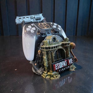 May include: A black and white PlayStation 5 controller sits on a detailed stand. The stand has a miniature scene with figures, a gun, and the words "Resident Evil Requiem."