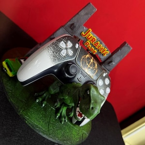 Könnte beinhalten: Ein individueller PlayStation 5-Controller mit Jurassic Park-Thema. Der Controller hat ein weißes und schwarzes Design mit einer goldenen Grafik. Eine grüne Dinosaurierfigur und das Jurassic Park-Logo sind ebenfalls vorhanden.