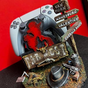 Op de afbeelding: Een decoratieve gamecontrollerstandaard met een Far West-thema. De standaard heeft een grijze controller, een rood cowboy-silhouet en een wegwijzer met plaatsnamen. Extra elementen zijn een hoed, geweren en een Far West-bord.