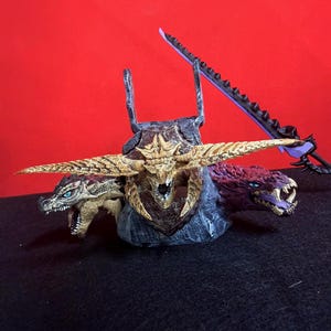 Könnte beinhalten: Detaillierte Fantasy-Figur mit einem dreiköpfigen Drachen in Gold- und Grautönen. Der Drache hat scharfe Zähne und blaue Augen. Ein lila Schwert mit gezackter Kante befindet sich hinter dem Drachen.