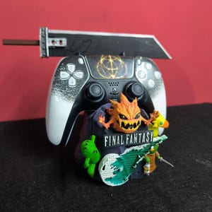Könnte beinhalten: Ein schwarz-weißer PlayStation 5-Controller mit einem dekorativen Ständer mit Final Fantasy-Charakteren und einem großen Schwert. Der Ständer enthält ein feuriges Monster, eine grüne Kreatur und einen gelben Vogel. Der Text "FINAL FANTASY" ist sichtbar.