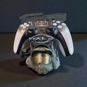 Könnte beinhalten: Ein schwarz-weißer PlayStation-Controller liegt auf einem 3D-gedruckten Ständer mit einem Halo-Helm und dem Wort "HALO" in Blau. Der Controller hat weiße Akzente und ein goldenes Design.