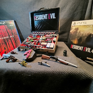May include: A collection of Resident Evil video game merchandise, including a suitcase with the game's logo, a PlayStation 2 game case, a PlayStation 5 game case, and various miniature weapons and accessories.