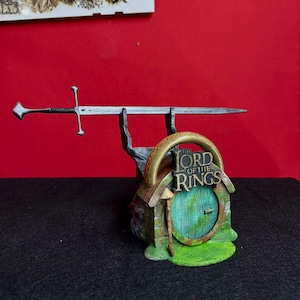 May include: A miniature replica of a sword and a hobbit hole from "The Lord of the Rings." The sword is silver with a cross-shaped hilt. The hobbit hole has a green door and the words "The Lord of the Rings" on top.