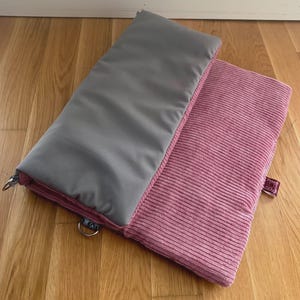 May include: Two rectangular pillows, one gray and one pink, rest on a wooden surface. The gray pillow has a smooth texture, while the pink one has a corduroy texture. Both pillows have a pink border. The pillows are likely for home decor or comfort.