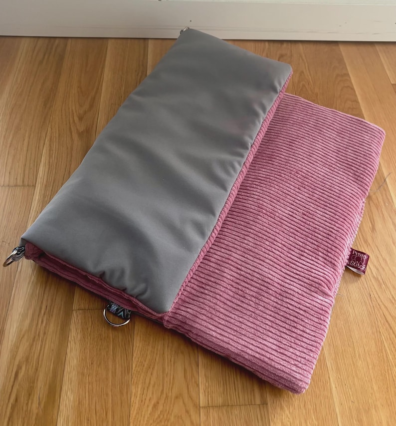 May include: Two rectangular pillows, one gray and one pink, rest on a wooden surface. The gray pillow has a smooth texture, while the pink one has a corduroy texture. Both pillows have a pink border. The pillows are likely for home decor or comfort.