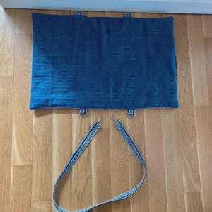 May include: A teal corduroy pet bed with metal D-rings and a patterned strap. The bed is rectangular and has a ribbed texture. The strap is blue and white with a chevron design. The bed is designed for pets.