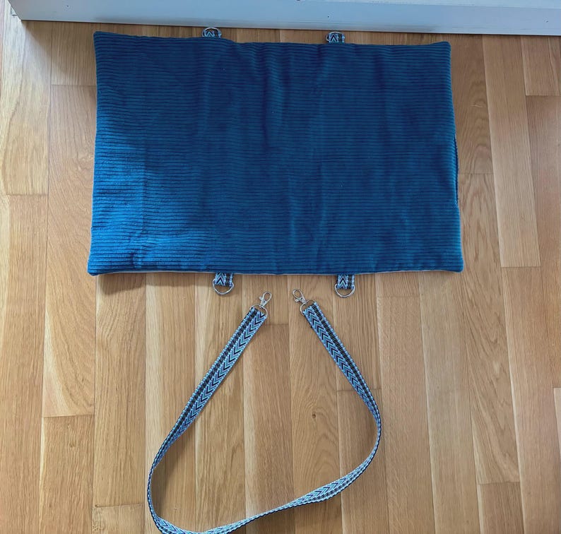 May include: A teal corduroy pet bed with metal D-rings and a patterned strap. The bed is rectangular and has a ribbed texture. The strap is blue and white with a chevron design. The bed is designed for pets.