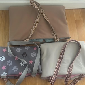 May include: Three pet carrier bags in different colors. One is tan with a patterned strap, another is gray with paw prints and pink stars, and the third is off-white with a patterned strap. All have straps for carrying.
