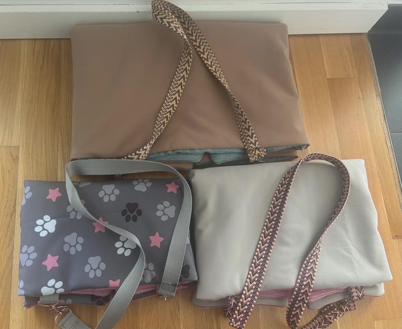 May include: Three pet carrier bags in different colors. One is tan with a patterned strap, another is gray with paw prints and pink stars, and the third is off-white with a patterned strap. All have straps for carrying.