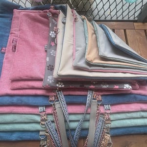 May include: A stack of pet bed mats in various colors, including blue, pink, and gray. Some mats have a corduroy texture, while others feature paw print patterns. The mats are rectangular and have attached straps.