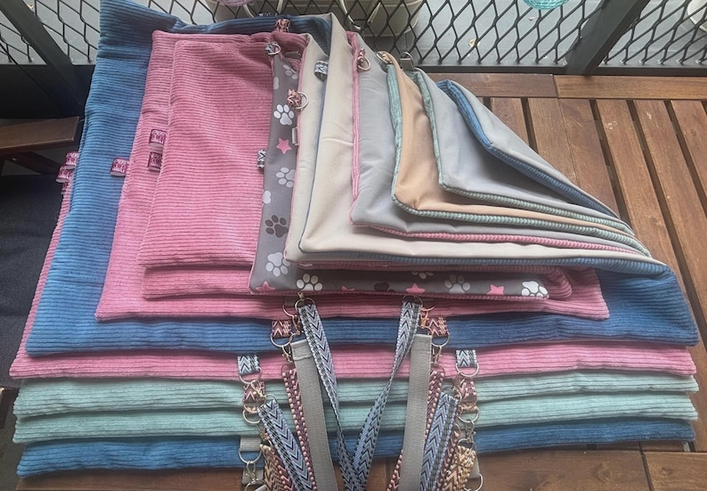 May include: A stack of pet bed mats in various colors, including blue, pink, and gray. Some mats have a corduroy texture, while others feature paw print patterns. The mats are rectangular and have attached straps.