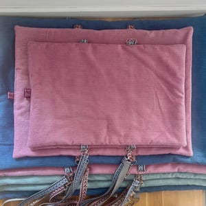 May include: A stack of corduroy pet beds in various sizes and colors. The beds are in shades of blue, pink, and teal. Each bed has a tag and a decorative strap with metal hardware.