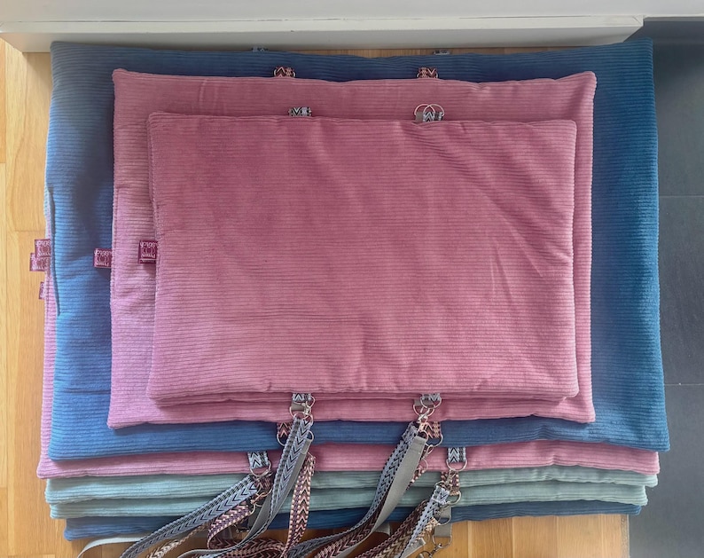 May include: A stack of corduroy pet beds in various sizes and colors. The beds are in shades of blue, pink, and teal. Each bed has a tag and a decorative strap with metal hardware.