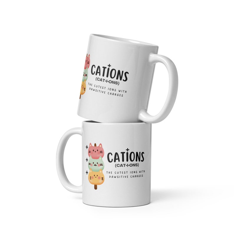 May include: Two white ceramic mugs stacked on top of each other. Both mugs feature a graphic of three cartoon cats stacked on top of each other, resembling a Japanese dessert. The text "CATIONS (CAT-I-ONS) THE CUTEST IONS WITH PAWSITIVE CHARGES" is printed on both mugs.