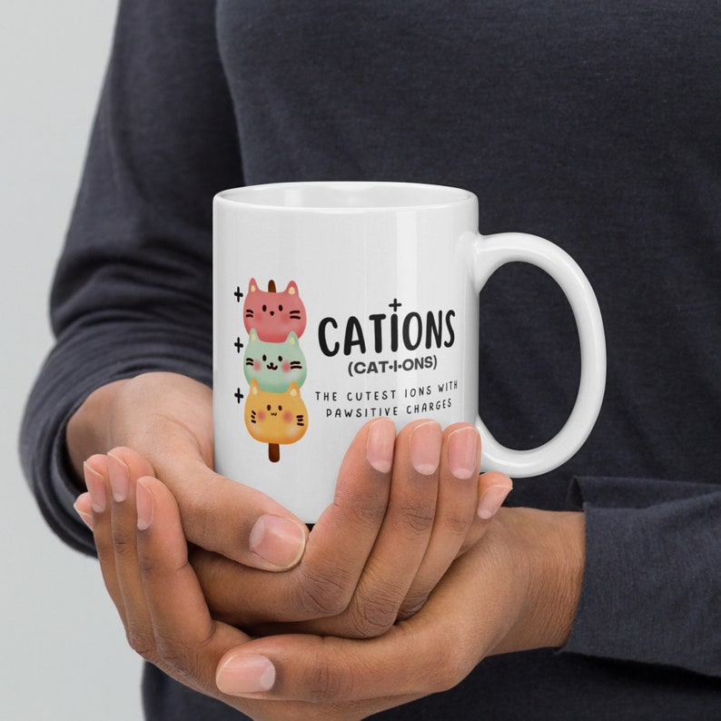May include: White ceramic mug with a black and white illustration of three cartoon cats stacked on top of each other. The text "CATIONS (CAT-I-ONS) THE CUTEST IONS WITH PAWSITIVE CHARGES" is printed on the mug.