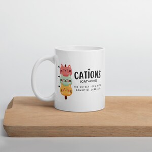 May include: White ceramic mug with a black and white illustration of three cartoon cats stacked on top of each other. The text "CATIONS (CAT-I-ONS) THE CUTEST IONS WITH PAWSITIVE CHARGES" is printed on the mug.