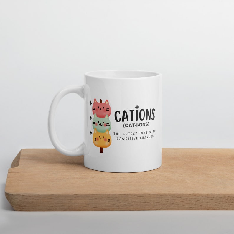 May include: White ceramic mug with a black and white illustration of three cartoon cats stacked on top of each other. The text "CATIONS (CAT-I-ONS) THE CUTEST IONS WITH PAWSITIVE CHARGES" is printed on the mug.