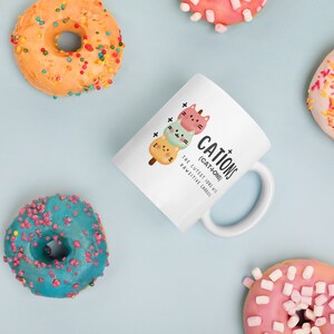 May include: White ceramic mug with a black and white illustration of three cartoon cats stacked on top of each other. The text "CATIONS (CAT-IONS) THE CUTEST IONS WITH PAWSITIVE CHARGES" is printed on the mug.