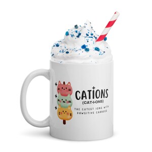 May include: White ceramic mug with a handle. The mug has a graphic of three cartoon cats stacked on top of each other. The text on the mug reads "CATIONS (CAT-I-ONS) THE CUTEST IONS WITH PAWSITIVE CHARGES".