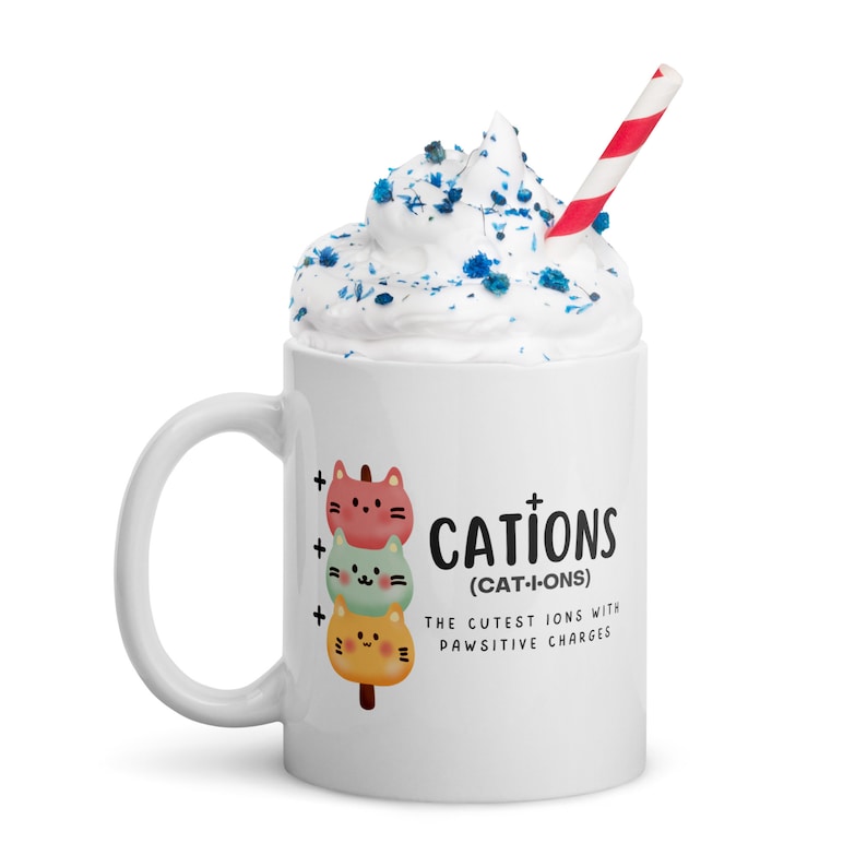 May include: White ceramic mug with a handle. The mug has a graphic of three cartoon cats stacked on top of each other. The text on the mug reads "CATIONS (CAT-I-ONS) THE CUTEST IONS WITH PAWSITIVE CHARGES".