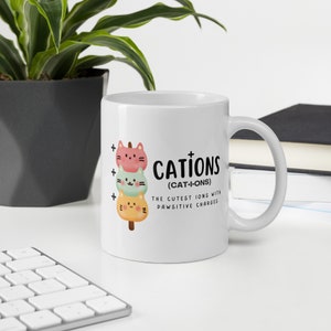 May include: White ceramic mug with a graphic of three stacked cartoon cats with plus signs next to them. The text reads "Cations (Cat-I-ons) The Cutest Ions With Pawsitive Charges."