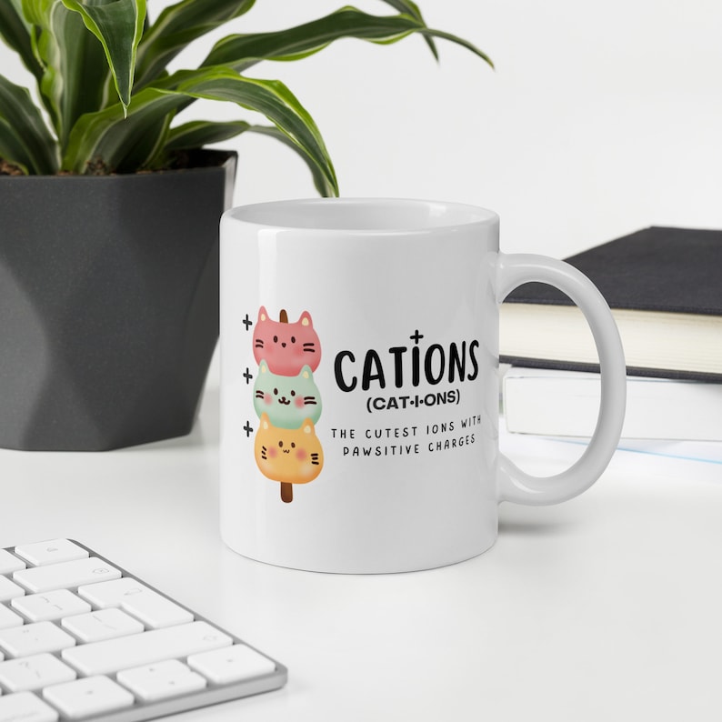 May include: White ceramic mug with a graphic of three stacked cartoon cats with plus signs next to them. The text reads "Cations (Cat-I-ons) The Cutest Ions With Pawsitive Charges."