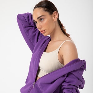 May include: A purple zip-up hoodie with a light beige tank top underneath. The hoodie has a zipper that is partially open.