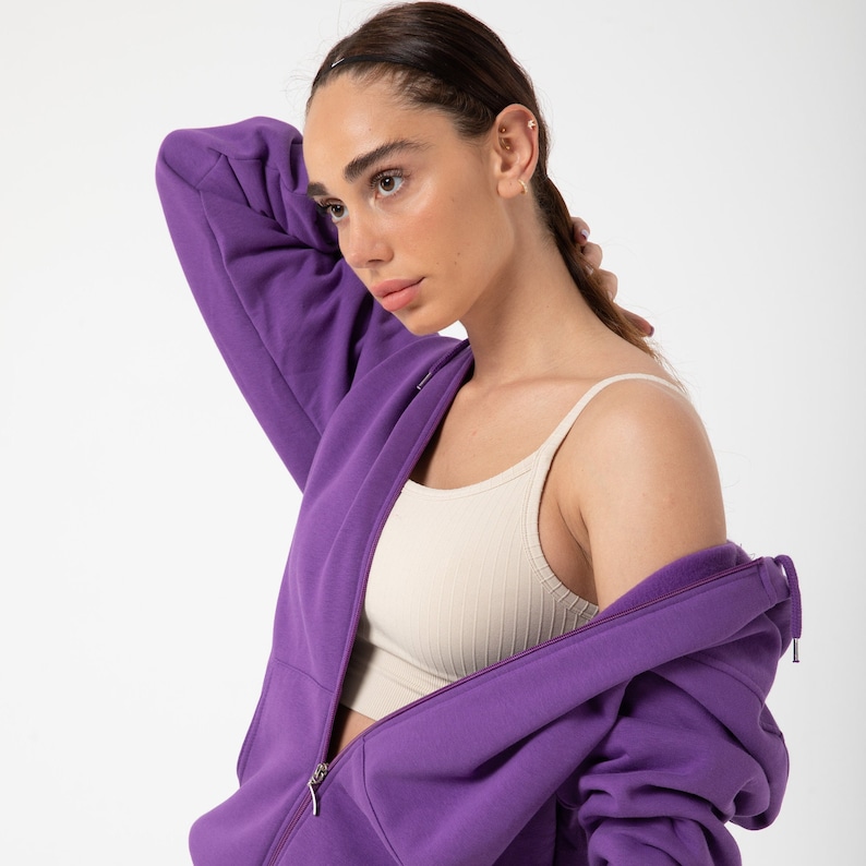 May include: A purple zip-up hoodie with a light beige tank top underneath. The hoodie has a zipper that is partially open.
