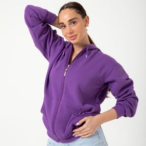 Puede incluir: Una sudadera con capucha morada con cremallera, capucha con cordón y bolsillos delanteros. La sudadera tiene una cremallera plateada y está hecha de un material suave y cómodo. Es una prenda de vestir informal.