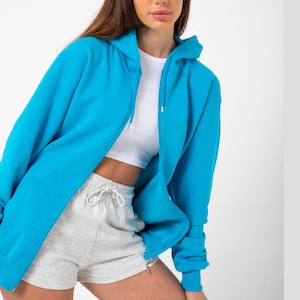 May include: A bright blue zip-up hoodie with a hood and long sleeves. The hoodie is worn over a white crop top and grey shorts.