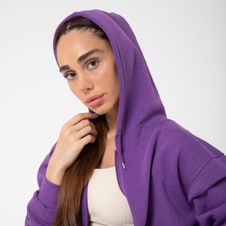 May include: A woman wearing a purple hooded sweatshirt with a zip. The sweatshirt has a relaxed fit and is made of a soft, comfortable fabric.