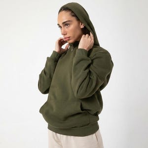 May include: An olive green hooded sweatshirt with a kangaroo pocket. The sweatshirt has a drawstring hood and long sleeves. The person is wearing beige pants. The background is plain white.