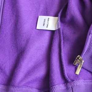 May include: A bright purple fleece-lined zip-up hoodie featuring a silver-toned metal zipper pull and a white interior garment tag that reads Made in Turkey and 100% Cotton. The fabric has a soft, fuzzy texture throughout the interior surface.
