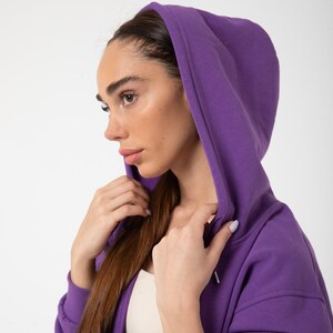 May include: A woman wearing a purple hooded sweatshirt with the hood pulled up over her head. She is looking to the side and holding the hood with her right hand.