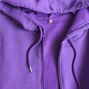 May include: A solid purple zip-up hoodie featuring a drawstring hood with matching purple cords and silver-toned metal aglets. The interior of the garment is lined with a soft fleece material, and a small white tag with the letter M is visible.