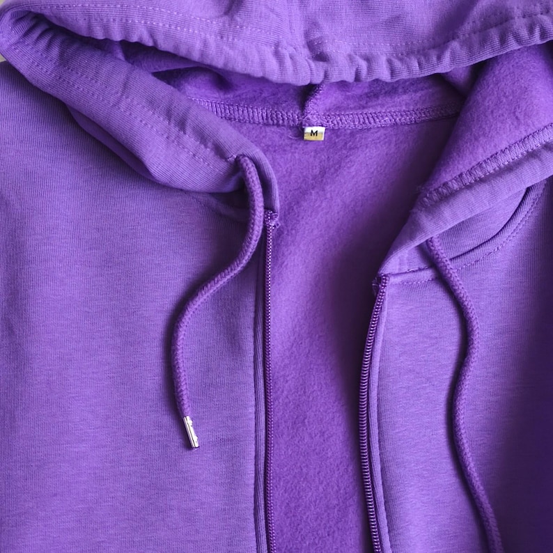 May include: A solid purple zip-up hoodie featuring a drawstring hood with matching purple cords and silver-toned metal aglets. The interior of the garment is lined with a soft fleece material, and a small white tag with the letter M is visible.