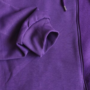 May include: A solid purple zip-up hoodie featuring a ribbed cuff on the sleeve and a matching purple zipper track. The fabric has a smooth, consistent texture throughout, with a drawstring cord visible near the top of the zipper closure.
