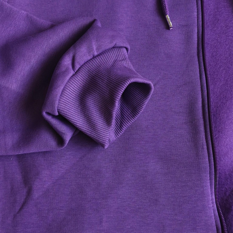 May include: A solid purple zip-up hoodie featuring a ribbed cuff on the sleeve and a matching purple zipper track. The fabric has a smooth, consistent texture throughout, with a drawstring cord visible near the top of the zipper closure.