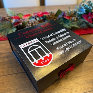 May include: Black gift box with a red ribbon. The box has a white label with the text "Connor Bell Class of 2024" and the University of Georgia logo. The text "School of Computing Bachelor of Science in Computer Science Minor in Japanese Language & Literature" is also on the label.